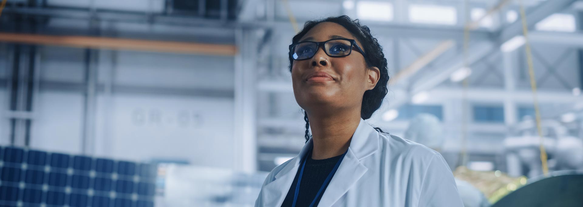 Black woman wearing glasses and a white lab coat inside a factory.